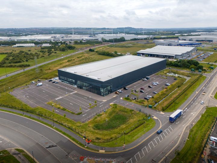 An aerial view of a facility with a parking lot, surrounded by a greenfield and a road