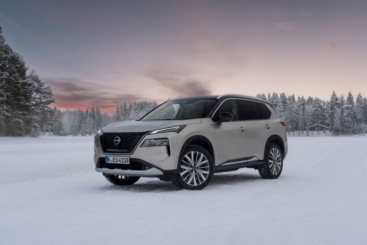 Silberner Nissan X‑Trail e‑Power e‑4ORcE steht auf schneebedecktem Eissee mit verschneiten Bäumen und Holzhütten im Hintergrund, Abendhimmel, 3/4 Front