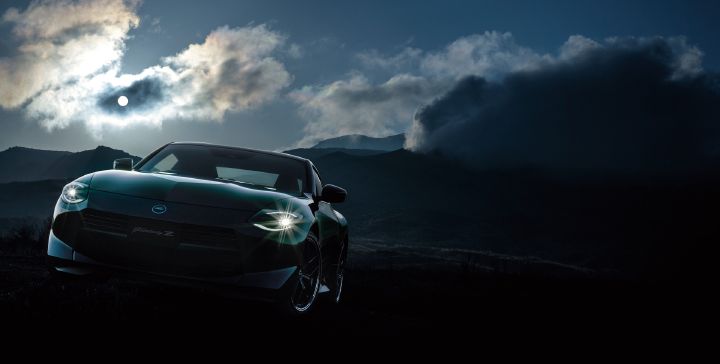 Front view of a Fairlady Z parked in a mountainous landscape at dawn or dusk. The vehicle faces directly toward the camera, with its headlights illuminated and shining brightly against the dark surroundings. Thick clouds fill the sky, while rays of light break through and illuminate the distant mountain ridges. The low-slung nose and wide stance emphasize the sports car’s dynamic character, creating a dramatic scene that highlights the Fairlady Z against the rugged natural backdrop.