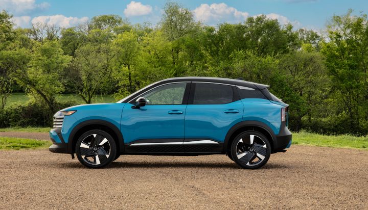 Left profile view of a blue 2025 Nissan Kicks parked on gravel with grass and lush foliage in the background.