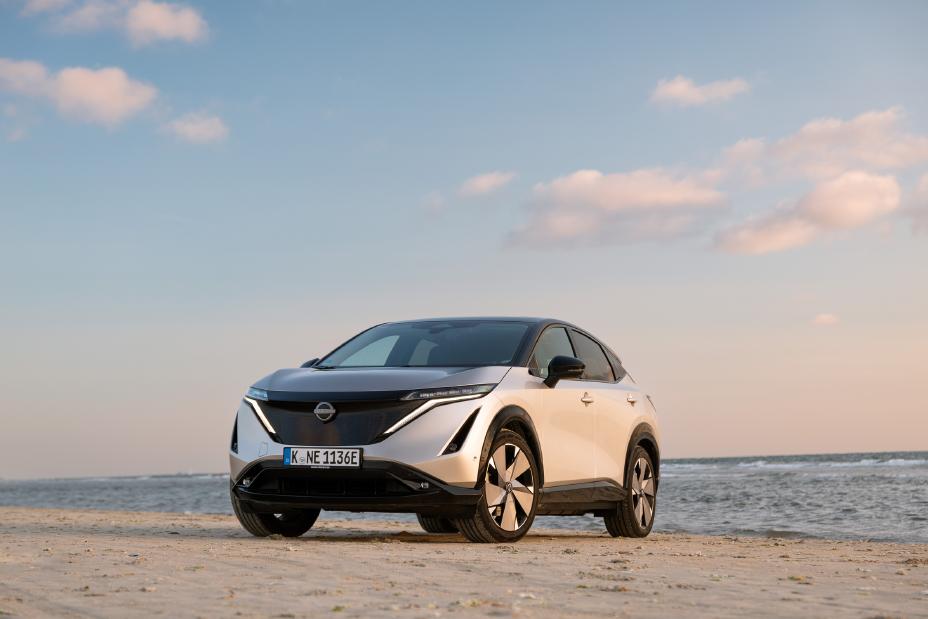 Ein silberner Nissan Ariya steht im Sand vor dem Meer, der blaue Himmel im Hintergrund färbt sich rosa