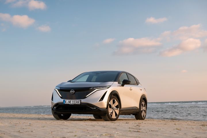 Ein silberner Nissan Ariya steht im Sand vor dem Meer, der blaue Himmel im Hintergrund färbt sich rosa
