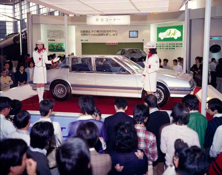 A crowded car exhibition displaying a light gray vehicle. 2 presenters dressed in white and red are addressing the crowd.