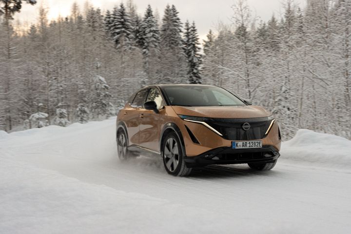 Elektroauto Nissan Ariya e‑4ORCE in Akatsuki Copper mit Dach und Außenspiegel in Pearl Black fährt auf schneebedeckter Straße mit verschneitem Wald im Hintergrund, 3/4 Front
