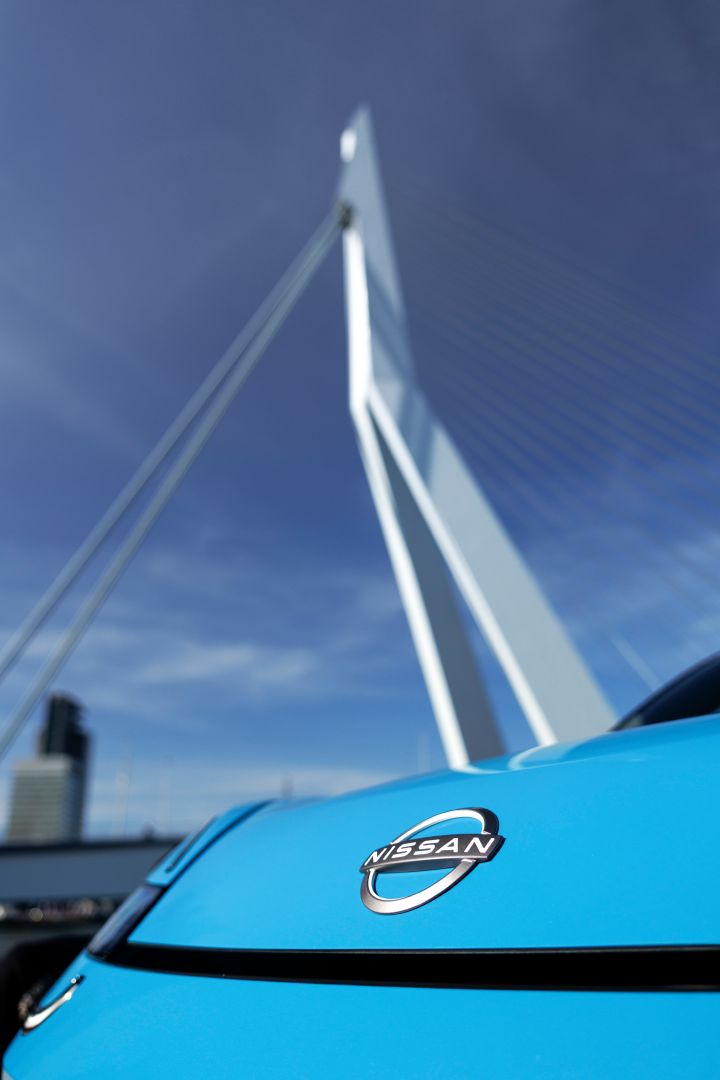 Detailaufnahme des Nissan Logos auf der Fronthaube eines blauen Nissan Micra, im Hintergrund sieht man unscharf eine Brücke mit hohem weißen Pfeiler in Rotterdam