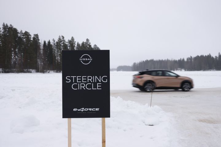 Schwarzes Schild mit weißer Schrift Nissan e‑4ORCE Steering Circle steht im Vordergrund im Schnee, Seitenansicht des im Hintergrund vorbeifahrenden Elektroautos Nissan Ariya e‑4ORCE in Akatsuki Copper mit Dach und Außenspiegel in Pearl Black