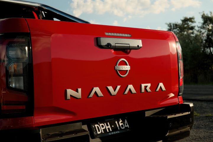 Close-up of the tailgate on a 2026 Nissan Navara ST-X in Outback Red, showing the Nissan badge, tailgate handle, number plate, and large NAVARA lettering between the rear lamp edges.