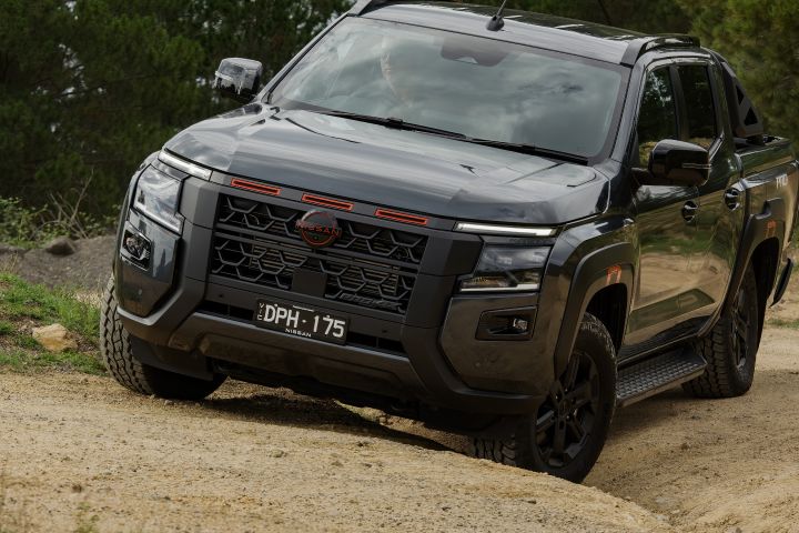 2026 Nissan Navara PRO-4X finished in Boulder Grey captured descending rutted sand from the front three‑quarter, showing red‑slatted grille, black bumper, side steps and front wheel articulation.