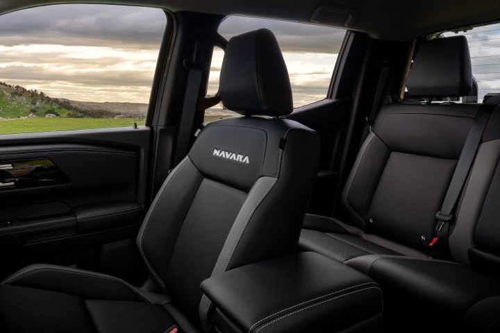 Interior seating view of a 2026 Nissan Navara ST-X showing the front seat with NAVARA embroidery, the centre armrest, the rear bench seat, and side windows with a rural landscape outside.