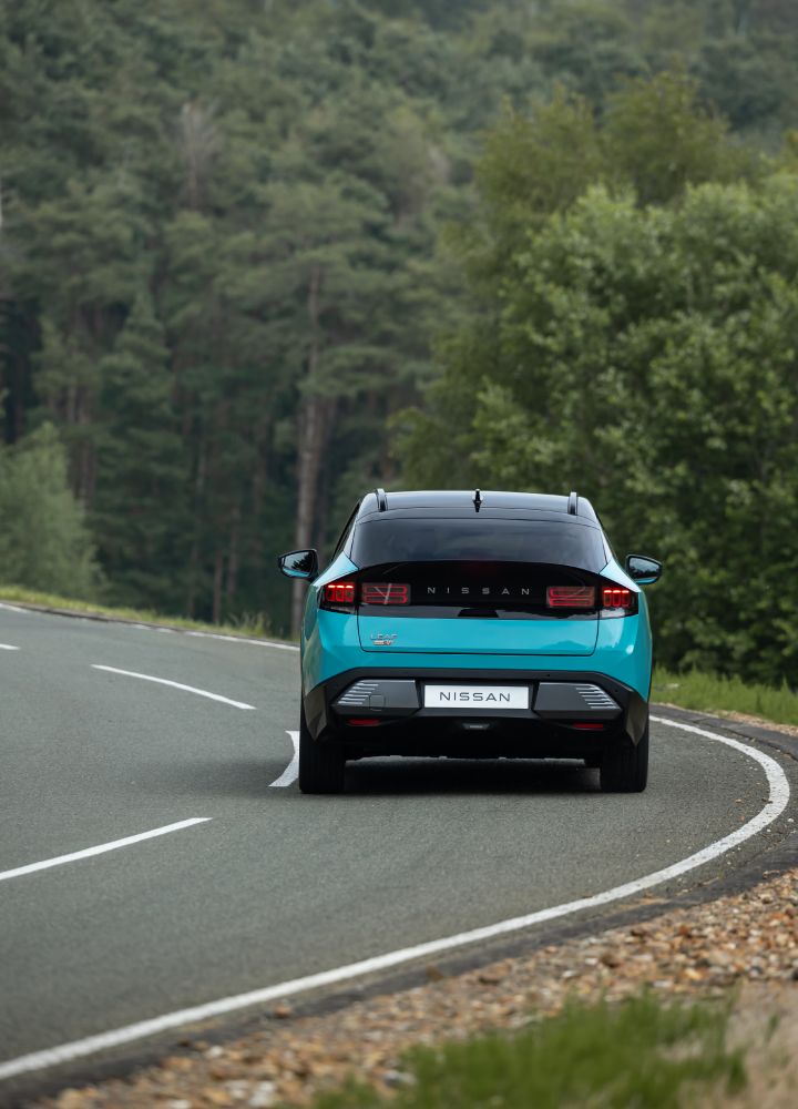 Vue arrière d'une Nissan LEAF bleu turquoise prenant le virage d'une route, avec de grands arbres verdoyants en arrière-plan