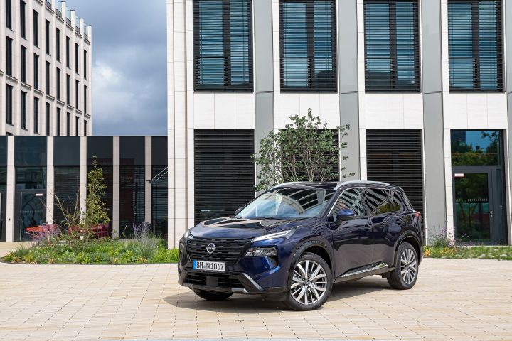 Blick von schräg vorne auf einen dunkelblauen Nissan X‑Trail, der auf einem hell gepflasterten Platz eines Bürogebäudes steht