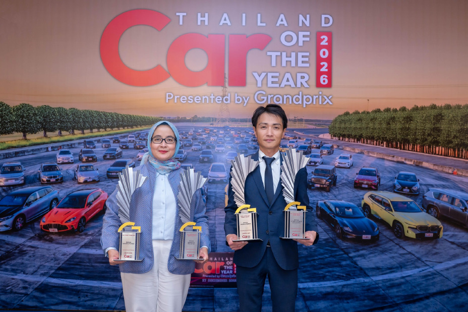 Two individuals stand in front of the “Thailand Car of the Year 2026” backdrop, each holding two trophies—one in each hand. The backdrop displays rows of cars on a test track with event branding at the top.