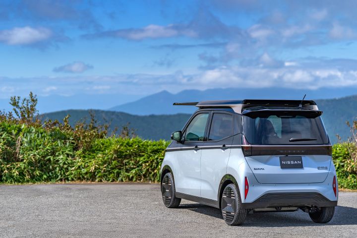 Against a backdrop of a lake and mountains, a light blue Nissan SAKURA is parked by the roadside, facing slightly left rear. The solar roof extends forward, with green shrubs nearby and distant blue mountains under a partly cloudy sky.