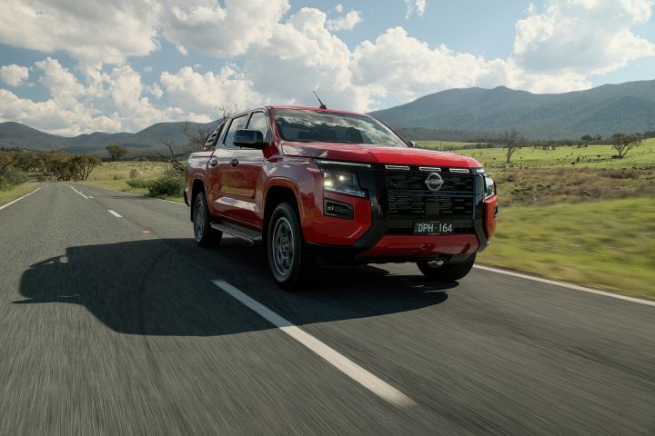 Front three‑quarter view of a 2026 Nissan Navara ST‑X dual cab in Outback Red, travelling on a sealed road past trees and rugged sloping terrain.