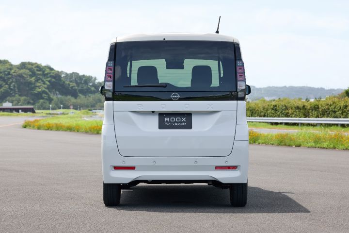 The new Nissan Roox kei car, in white, is shown from the rear, parked on a paved road with a backdrop of lush green trees and a blue sky.