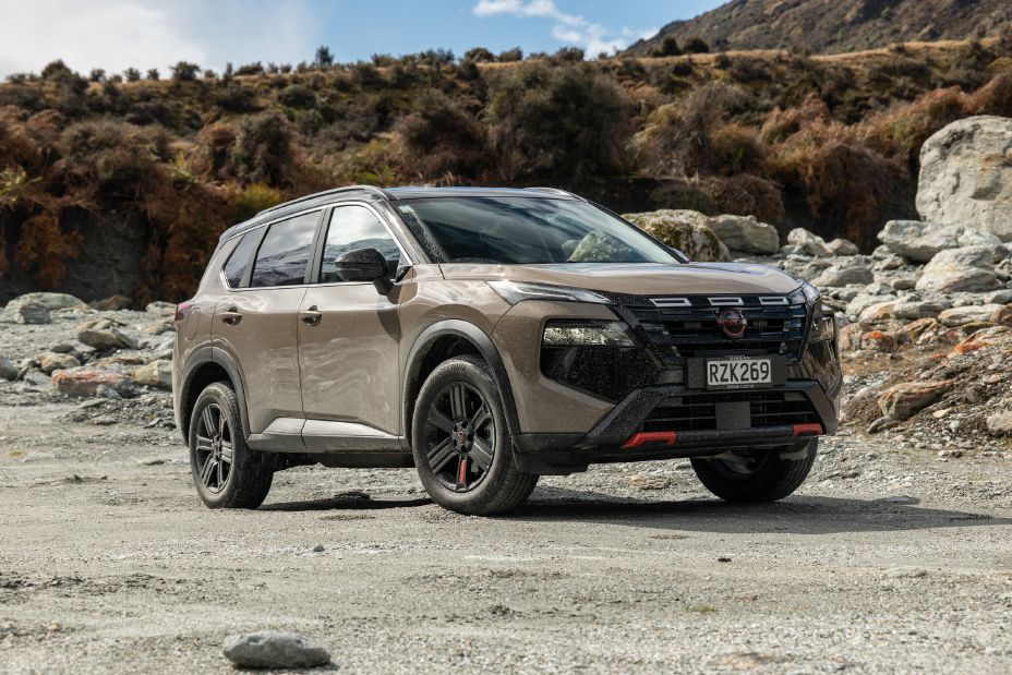 Close front three-quarter view of a 2026 Nissan X‑TRAIL Rock Creek in Baja Storm on rocky alpine terrain, showcasing its rugged grille, black alloy wheels and red exterior accents.