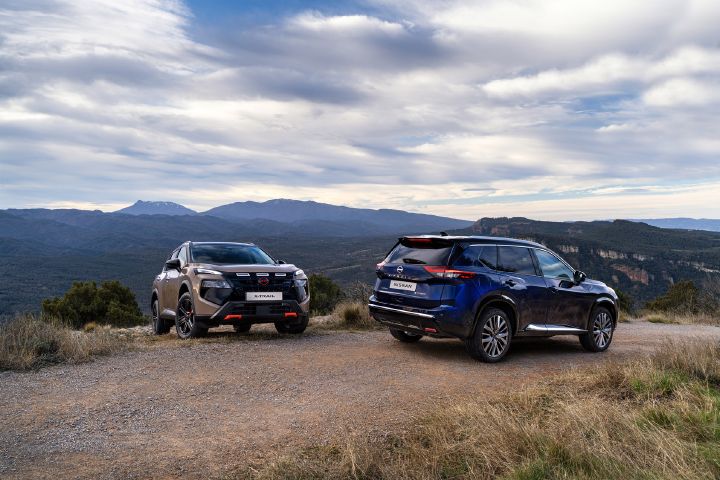 Twee nieuwe Nissan X‑Trail SUV’s uit 2026, geparkeerd op een onverharde open plek met uitzicht op een groot meer, omgeven door beboste heuvels en bergen onder een bewolkte hemel. Eén N-Trek in Baja Storm en één Tekna in Universal Blue zijn zo opgesteld dat zowel de voor- als achterzijde zichtbaar zijn, waarmee het design en de terreincapaciteiten in een schilderachtige omgeving worden benadrukt.
