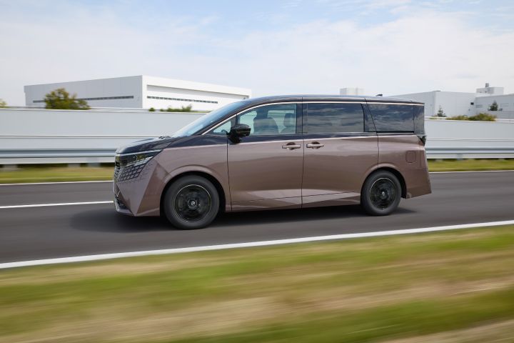 新型エルグランドが、画面右から左へ向かって直線道路を走行する側面ビューで捉えられている。車両は左方向へ進行しており、低めの視点からその伸びやかなボディラインとブラックルーフ、ダークトーンのホイールが強調されている。背景には白い工場の建物やガードレール、淡い青空と薄い雲が広がり、道路脇の芝生や周囲はモーションブラーで流れるように描かれ、走行中のスピード感が表現されている。