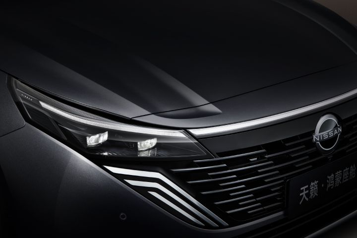 Close-up of the left front side of a black Teana, highlighting the sharp LED headlights and the grille with distinctive horizontal lines. The body has a metallic finish with a glossy texture.