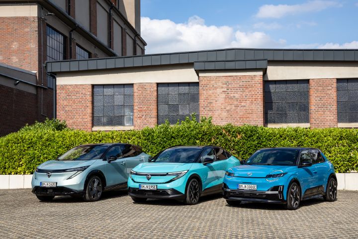 Vor einem Backsteingebäude und einer grünen Hecke stehen nebeneinander die drei Nissan EV-Modelle Nissan Ariya, Nissan Leaf und Nissan Micra