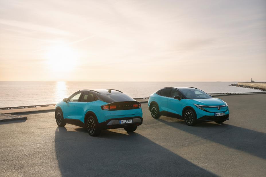 Two turquoise Nissan LEAF vehicles parked on a waterfront road at sunset, with the ocean and bright sun in the background.