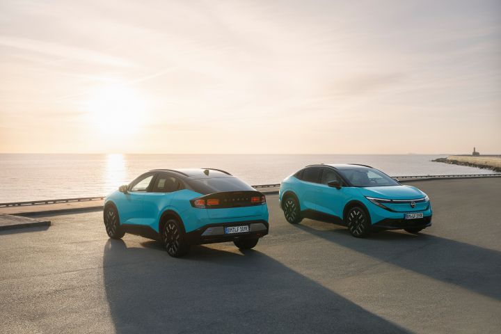 Two turquoise Nissan LEAF vehicles parked on a waterfront road at sunset, with the ocean and bright sun in the background.