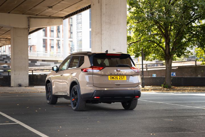 Rear view of a champagne-coloured Nissan X‑Trail parked in an urban setting, with trees, buildings and an overpass in the background