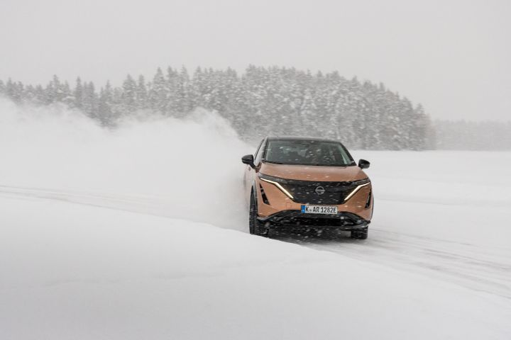 Elektroauto Nissan Ariya e‑4ORCE in Akatsuki Copper mit Dach und Außenspiegel in Pearl Black driftet im Schnee auf Eissee mit schneebedeckten Bäumen im Hintergrund, Front