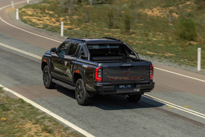 2026 Nissan Navara PRO-4X finished in Boulder Grey seen from the rear three‑quarter while driving, with NAVARA‑stamped tailgate, vertical LED tail‑lamps, sports bar and tow hitch on country bitumen.