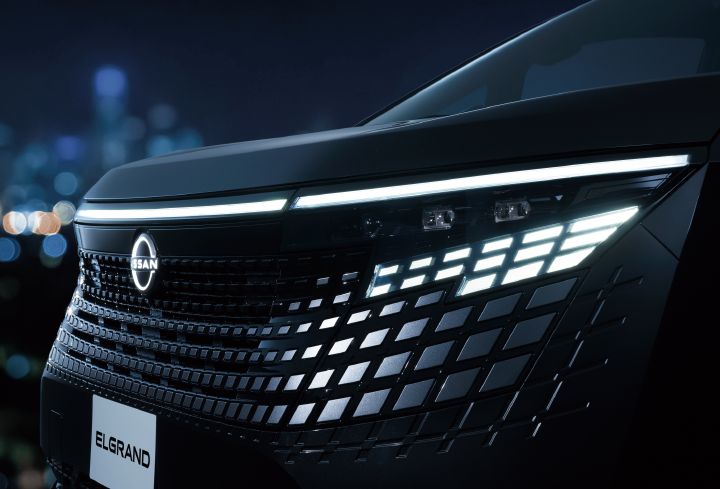 ブラックのエルグランド VIPのフロントフェイス右側を、車両前方やや左側からクローズアップで撮影した画像。車両は左方向を向いており、画面中央左には日産エンブレムを備えた大型フロントグリルが配置されている。グリルの下部とヘッドライトの下にはダークメタルグレーの加飾が配され、、ブラック基調の細かな矩形パターンが幾重にも並び、光を受けて立体的な表情を見せている。
画面右上には薄型のLEDヘッドランプが配置され、その下には複数の矩形発光ユニットで構成された特徴的なLEDシグネチャーランプが白く輝いている。ボディカラーは深みのあるブラックで、ヘッドランプ周辺の光沢のあるブラックパネルやグリル加飾と一体感のあるデザインとなっている。