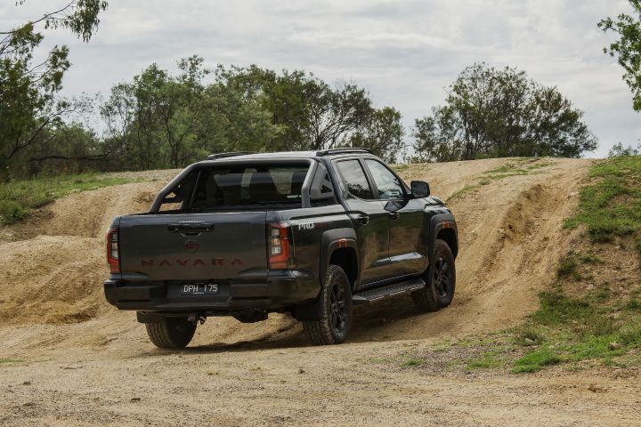 2026 Nissan Navara PRO-4X finished in Boulder Grey pictured rear three‑quarter departing along a sandy track with side steps, wheel‑arch cladding and receding NAVARA script.