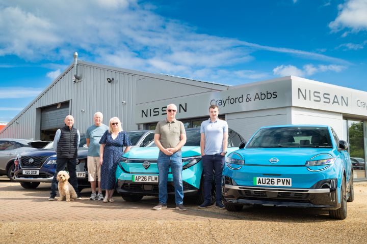 Five people and a dog standing in front of a Nissan X‑Trail, Ariya, LEAF and MICRA outside a Nissan dealership