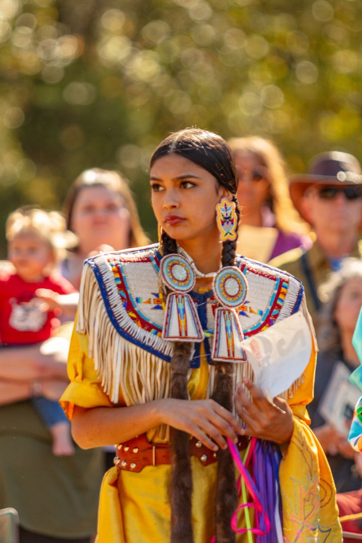 A young Native American women dressed in her culture’s attire with long braids stands in front of a group of people.