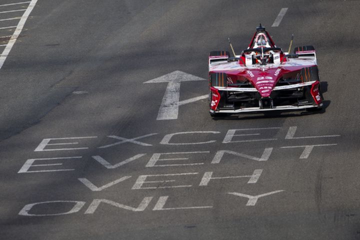 Ein rot-weißer Nissan Formel-E-Rennwagen fährt auf einer Fahrbahn vorwärts. Auf dem Asphalt sind große Richtungspfeile und die Aufschrift „EXCEL EVENT VEH ONLY“ aufgemalt, und das Fahrzeug ist von einer breiten, asphaltierten Straße umgeben.
