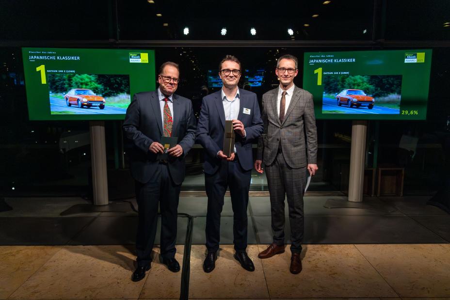 Drei Männer im Anzug stehen vor zwei Bildschirmen, Vincent Ricoux in der Mitte nimmt den Motor Klassik Award entgegen von Martin Puthz auf der linken Seite und Andreas Of-Allinger rechts.