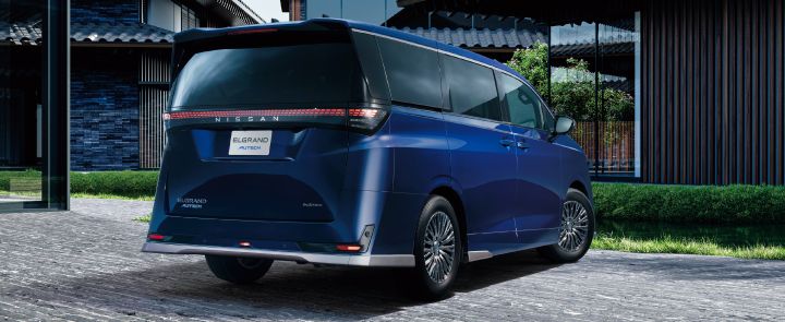 ブルーのエルグランド AUTECHを、左後方から撮影したリア右向きの3/4アングルの画像。車両は画面中央に配置され、フロント部分は右方向を向いている。リアには横一文字に伸びる赤色のコンビネーションランプが点灯し、中央に「NISSAN」の文字が配置されている。テールゲートには「ELGRAND」「AUTECH」「e‑4ORCE」のエンブレムが見え、バンパー下部にはメタル調フィニッシュの加飾が施されている。深いブルーのボディとダークカラーのウインドウ、精巧なデザインのアルミホイールが上質な印象を与えている。