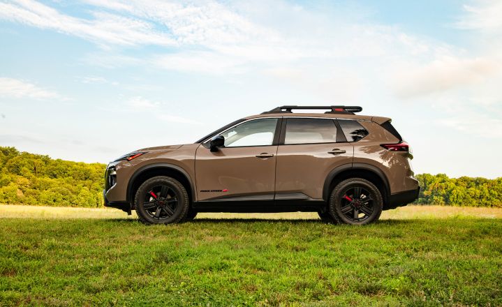 Left profile angle of a 2025 Nissan Rogue Rock creek parked on a pasture with trees and, blue skies around.