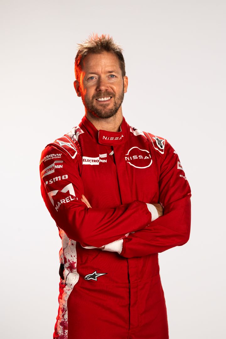 Sam Bird lächelt mit verschränkten Armen in seinem roten Rennanzug des Nissan Formula E Teams vor einem weißen Hintergrund.