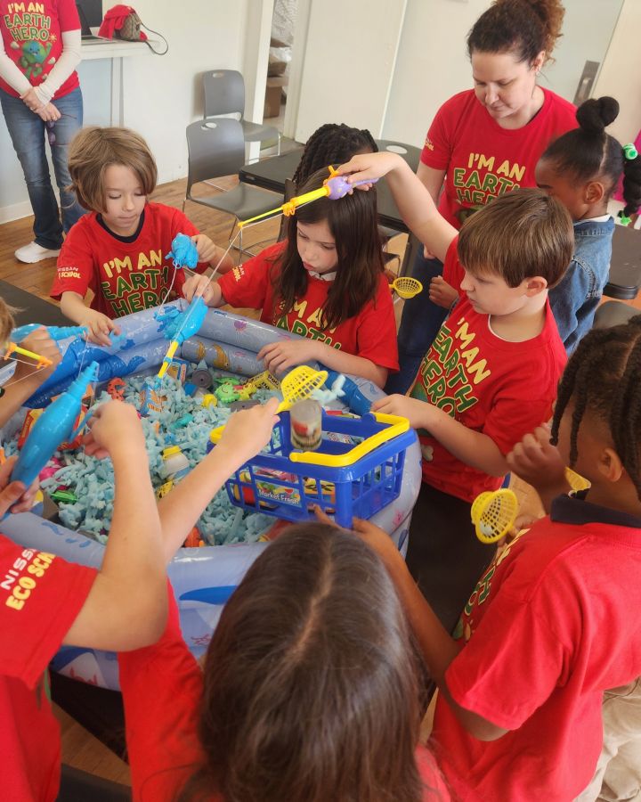 A group of children stand over a play fishing pool filled with foam pieces and toys.