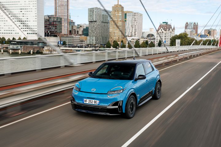 Ein blauer Nissan Micra fährt über eine Brücke in Amsterdam, im Hintergrund sind mehrere Hochhäuser zu sehen