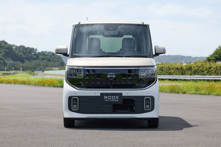 The new Nissan Roox kei car, in white, is parked facing forward on a paved road. The vehicle features a boxy design with a prominent grille, rectangular headlights, and side mirrors extending from the sides. The background includes green trees and foliage.