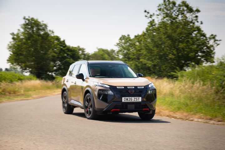 Champagne-coloured Nissan X‑Trail driving along a country road, viewed from the front three-quarter angle, with trees and grassland in the background.