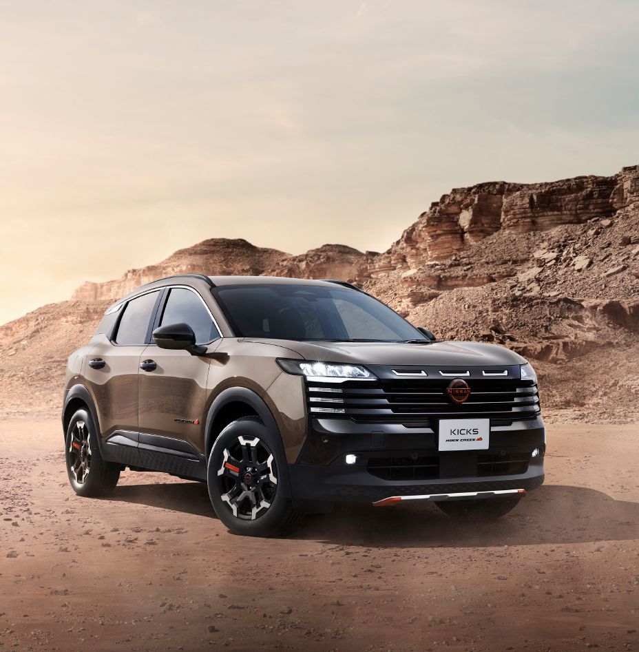 A kicks rock creek in a beige-toned body color is parked in a front three-quarter view facing right. The black-accented front grille, unique alloy wheels, and lava red accent parts create a rugged and powerful appearance. In the background, barren rocky mountains and cliffs with layered rock formations stretch across the landscape, while dry terrain covered with sand and small stones extends into the distance. Illuminated by natural light, the vehicle's tough suv styling stands out in this off-road environment.
