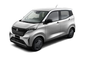 シルバーの「日産サクラ」を、無地の白背景の前で車両の左前方から斜めに撮影した画像。落ち着いた色合いのボディとフロントマスクが中央に写っている。