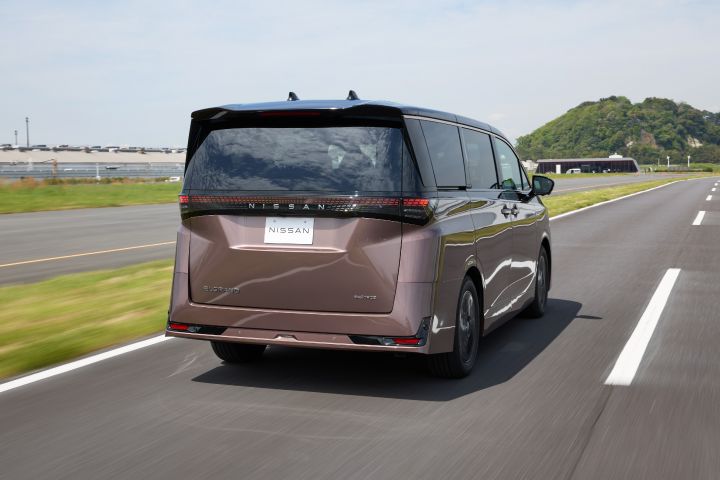 新型エルグランドが、右へ向かって直線道路を走行している後方斜めのアングルで捉えられている。車両は右方向へ進行しており、ブラックのルーフと一体化したリアウインドウ、横一文字に伸びたLEDテールランプ、中央の「NISSAN」ロゴが強調されている。背景には青空と薄い雲、緑の芝生や丘、小規模な建物が広がり、路肩や周囲はモーションブラーで流れるように描かれ、走行中のスピード感が表現されている。
