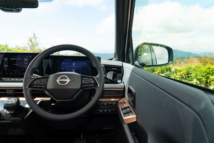 Interior view of a Nissan SAKURA showing a black steering wheel and digital display in the center. A gear selector is on the left, and the right side shows the door and mirror. Outside the window are green shrubs, mountains, and a blue sky.