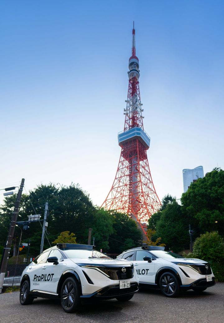 Zwei weiße Nissan Ariya Fahrzeuge stehen mit der Front nach vorne ausgerichtet in einer Reihe. Im Hintergrund ragt der Tokyo Tower unter einem strahlend blauen Himmel über üppig grünen Bäumen empor.