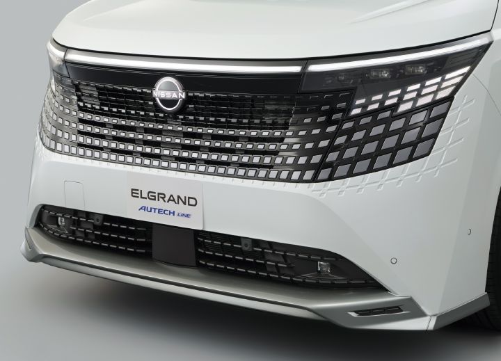ホワイトのエルグランド AUTECH LINEのフロント部分を、車両前方やや左側からクローズアップで撮影した画像。車両は右方向を向いており、画面中央には日産エンブレムを配した大型フロントグリルが配置されている。グリルは上部がブラック、下部がタークメタルグレーとされ、横方向に並ぶ細かな矩形パターンが立体的にデザインされている。
画面右上には薄型LEDヘッドランプが備わり、その下には幾何学模様のLEDシグネチャーランプが点灯している。フロントバンパー下部にはダークメタルグレーのスポイラー加飾が組み合わされ、上質かつ先進的な印象を与えている。
ボディカラーは光沢のあるホワイトで、ブラックのグリルやヘッドランプとのコントラストが際立っている。