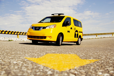 Un taxi Nissan NV200 amarillo fotografiado con una flecha amarilla en el piso de concreto