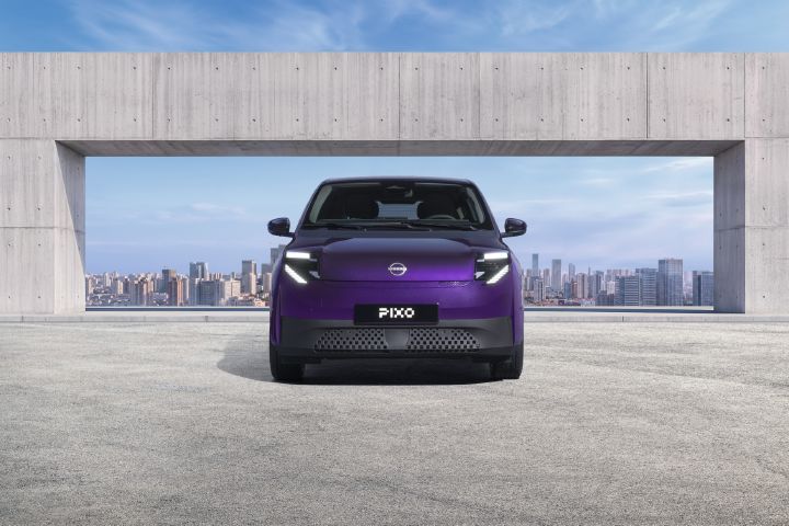 Foto, das ein violettes Nissan-Pixo-Konzeptfahrzeug zeigt, das vor einer architektonischen Öffnung aus Beton geparkt ist, hinter der sich die Skyline der Stadt abzeichnet. Die Vorderansicht hebt die beleuchteten, kantigen Scheinwerfer, den geschlossenen Kühlergrill, das Pixo-Nummernschild, das schnittige, kompakte Design und den blauen Himmel im Hintergrund hervor.